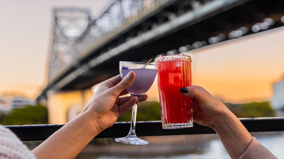 Two cocktails touching over the rail at Fiume, Story Bridge and golden sky behind