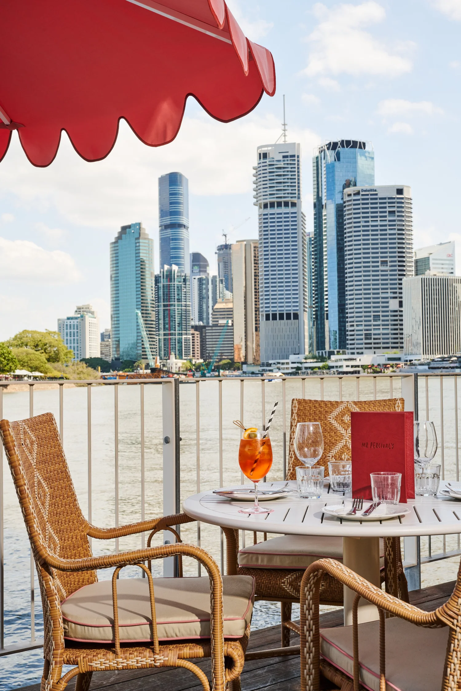 A river-edge table at Mr Percival's under a red scalloped umbrella, Brisbane towers across the water